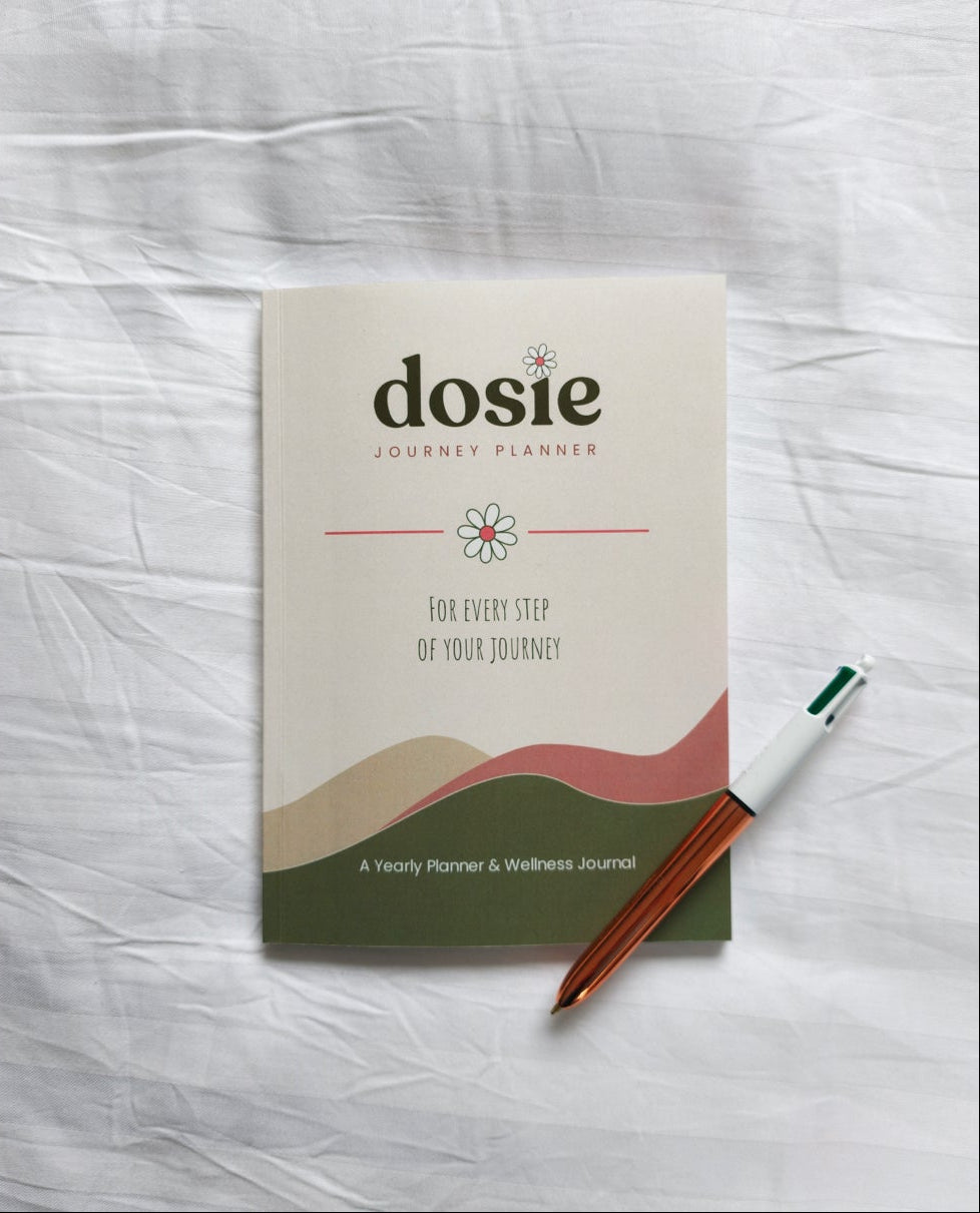 A dosie Journey Planner booklet lies on a wrinkled white fabric surface, with a brown pen placed beside it.